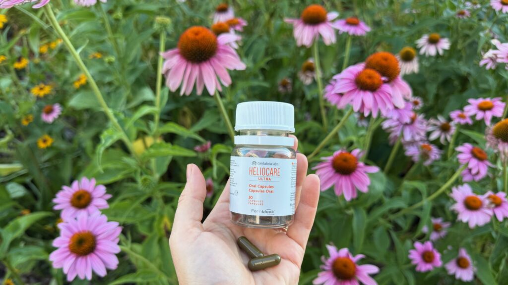 Fotografie originală cu suplimentul Heliocare Ultra Oral Capsules, flacon ținut în mână pe fundal de flori roz de echinacea.