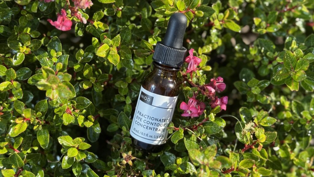 Sticluță de serum NIOD Fractionated Eye Contour Concentrate pentru îngrijirea pielii din jurul ochilor, fotografiată pe fundal natural cu frunze și flori.