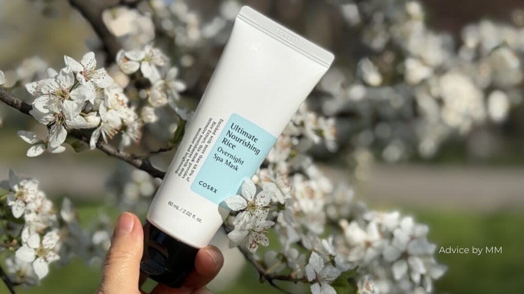 COSRX Ultimate Nourishing Rice Overnight Spa Mask ținut în mână, fotografiat în lumină naturală pe fundal de flori albe.