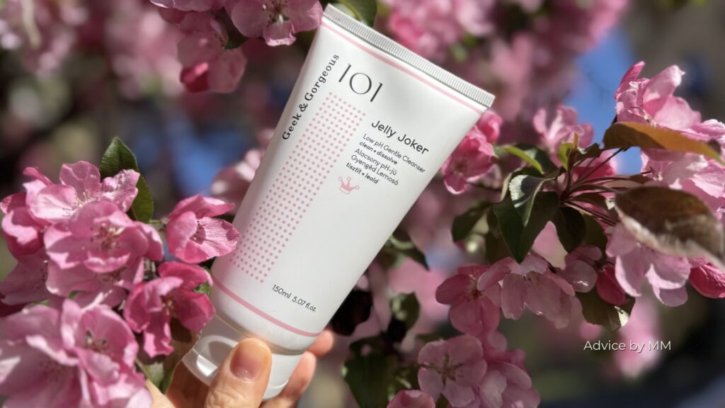 Geek & Gorgeous Jelly Joker, cleanser delicat cu pH scăzut, fotografiat printre flori roz; formulă conservată cu phenoxyethanol.