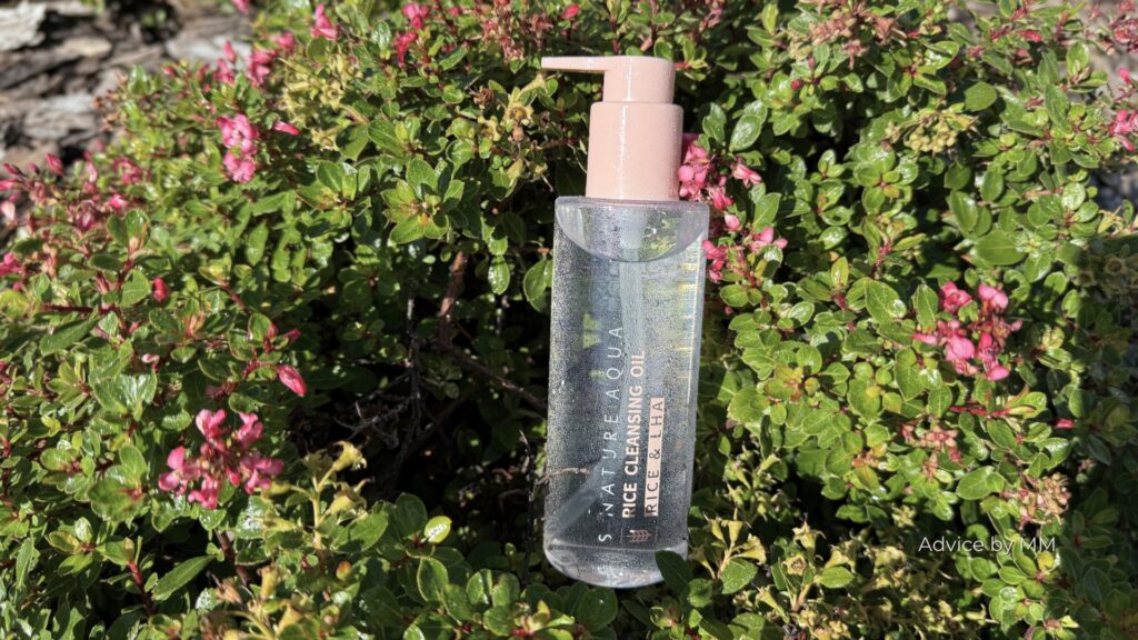S.NATURE Aqua Rice Cleansing Oil fotografiat între frunze verzi și flori roz, într-o imagine dedicată produselor de curățare cu orez.
