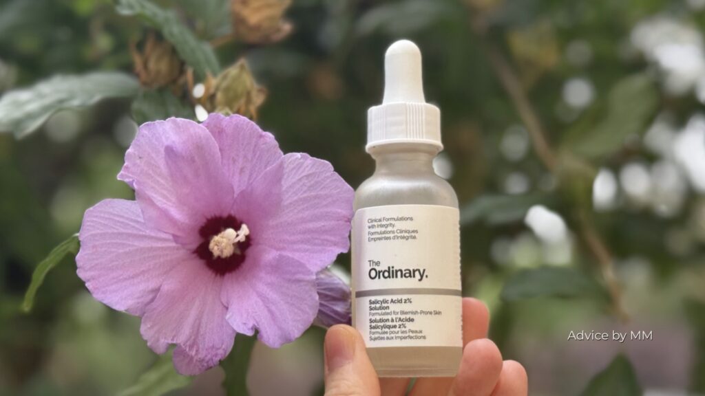 Flacon The Ordinary Salicylic Acid 2% Solution fotografiat în fața unei flori roz de hibiscus; produs conservat cu phenoxyethanol.