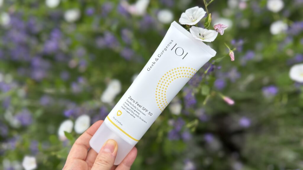 Tub de Geek & Gorgeous Zero Feel SPF 50 ținut în mână, fotografiat în aer liber pe fundal blurat de flori albe și mov. Produs de protecție solară utilizat ca exemplu într-un articol despre phenoxyethanol în cosmetice.